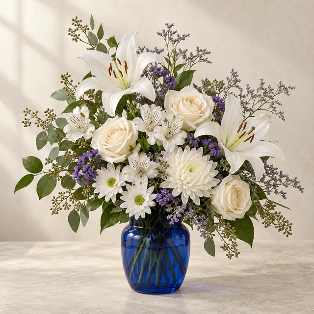 White lilies, cream roses, white chrysanthemums and alstroemeria with violet accents in a rounded cobalt-blue glass vase.