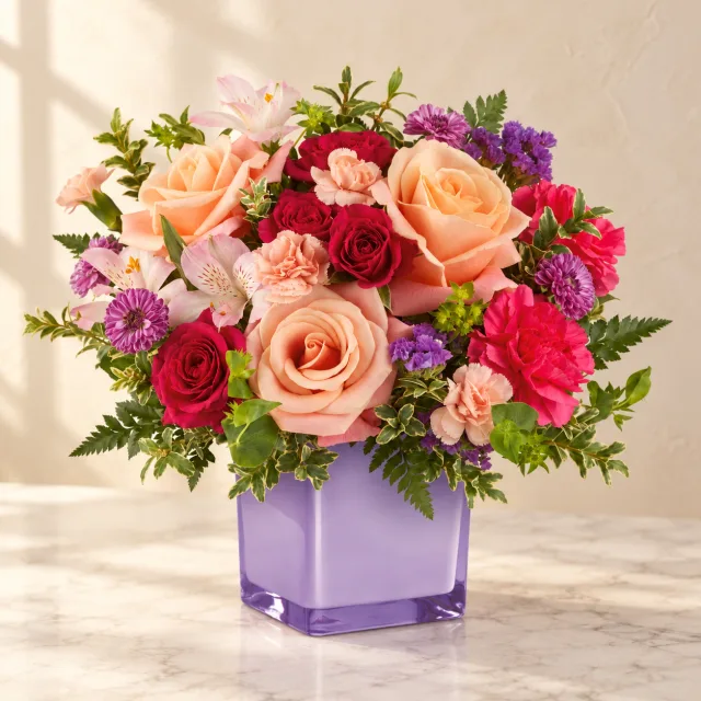 Peach roses, hot pink spray roses and pink alstroemeria in a lilac glass cube