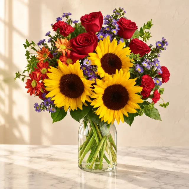 Sunflowers, red roses, bronze daisies and lavender asters in a round clear glass vase